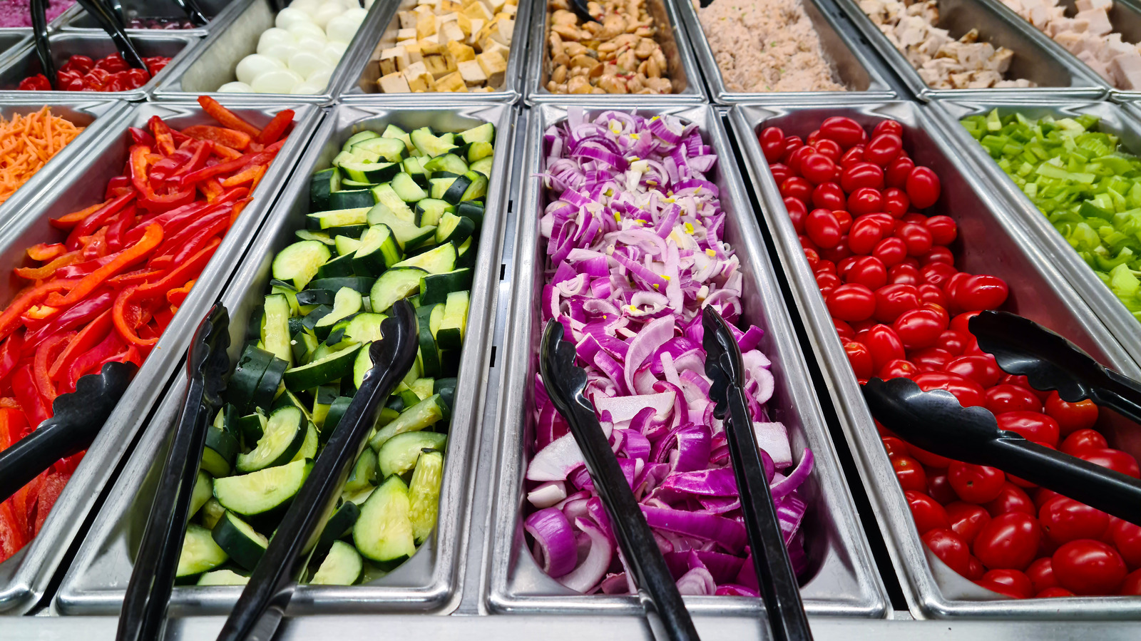 Stainless steel salad bar trays with bell peppers, cucumbers, cherry tomatoes, carrots, eggs, and serving tongs